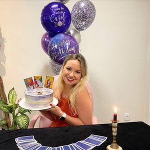 May include: A birthday cake decorated with tarot cards and a candle. The cake is on a white plate and there are purple and silver balloons in the background. The text on the balloons reads "Happy Birthday" and "2 anos de Tar&ocirc; da Carol".