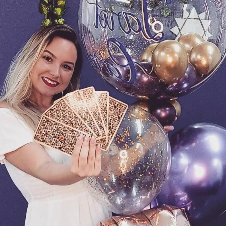 May include: A woman holds a fan of playing cards in front of a background of colorful balloons. The balloons are clear, purple, and gold. The clear balloons have glitter inside. The text "Happy Birthday" is written on one of the balloons.