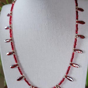 May include: A red beaded necklace with teardrop-shaped accents. The necklace features small red beads and larger, iridescent teardrop-shaped beads. The necklace is displayed on a white mannequin, showcasing its design and color.