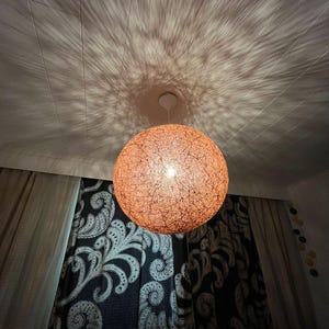 Op de afbeelding: Een bolvormige, oranje hanglamp van geweven materiaal hangt aan het plafond. Het licht werpt een sterrenpatroon op het plafond. Donkere gordijnen met een wit paisley-ontwerp zijn zichtbaar op de achtergrond.