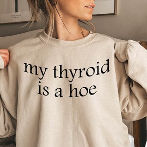 May include: A beige sweatshirt with the text "my thyroid is a hoe" printed in black.
