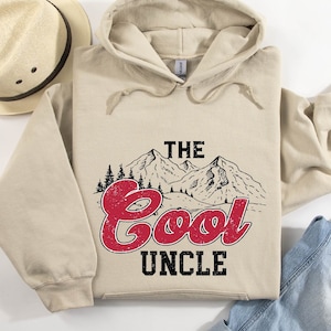 May include: A beige hoodie with the text "THE COOL UNCLE" in red and black, featuring a mountain and forest graphic. A straw hat is visible in the upper left corner, and blue jeans are in the lower right corner.