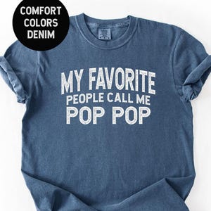 May include: Denim blue t-shirt with the text "MY FAVORITE PEOPLE CALL ME POP POP" in white. A black circle in the upper left corner reads "COMFORT COLORS DENIM". The shirt has rolled-up sleeves.