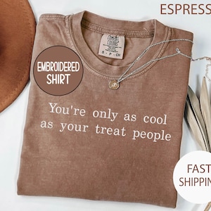 May include: A brown short-sleeved t-shirt with a crew neckline and a Comfort Colors label. The shirt features white embroidered text that reads, "You're only as cool as your treat people." A delicate silver-toned necklace rests on the fabric.