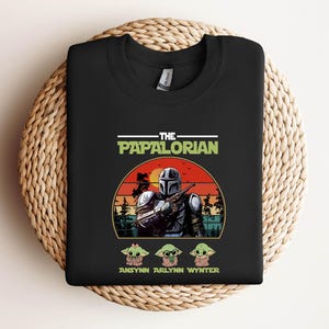 Puede incluir: Sudadera negra de cuello redondo con un gr&aacute;fico de un personaje al estilo Mandaloriano y el texto "THE PAPALORIAN". Debajo del gr&aacute;fico hay tres personajes de dibujos animados y los nombres "ANSYNN ARLYNN WYNTER."