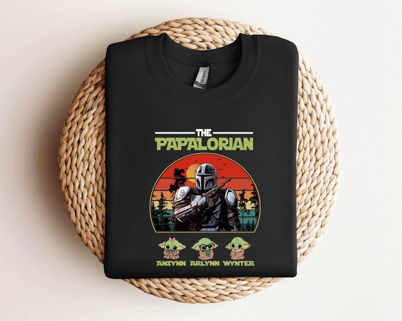 Puede incluir: Sudadera negra de cuello redondo con un gr&aacute;fico de un personaje al estilo Mandaloriano y el texto "THE PAPALORIAN". Debajo del gr&aacute;fico hay tres personajes de dibujos animados y los nombres "ANSYNN ARLYNN WYNTER."