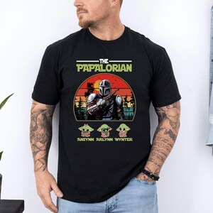 Puede incluir: Camiseta negra con un gr&aacute;fico de un personaje al estilo Mandaloriano, el texto "THE PAPALORIAN" y tres personajes Grogu. El dise&ntilde;o incluye un fondo de puesta de sol y los nombres "ANSYNN ARLYNN WYNTER".
