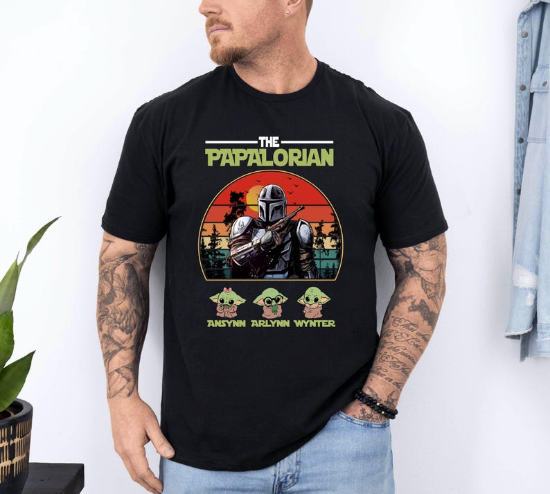 Puede incluir: Camiseta negra con un gr&aacute;fico de un personaje al estilo Mandaloriano, el texto "THE PAPALORIAN" y tres personajes Grogu. El dise&ntilde;o incluye un fondo de puesta de sol y los nombres "ANSYNN ARLYNN WYNTER".