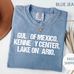 May include: A blue short-sleeve t-shirt with a distressed white graphic print that reads GULF OF MEXICO. KENNEDY CENTER. LAKE ONTARIO. The shirt features a crew neckline and a Comfort Colors brand label at the inner collar, paired with a gold necklace.
