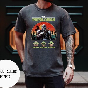Puede incluir: Camiseta gris oscuro con un gr&aacute;fico que presenta un personaje al estilo Mandaloriano y el texto "THE PAPALORIAN". Debajo del gr&aacute;fico hay tres personajes de Grogu y los nombres "ANSYNN ARLYNN WYNTER". La camiseta tambi&eacute;n tiene el texto "COMFORT COLORS PEPPER".