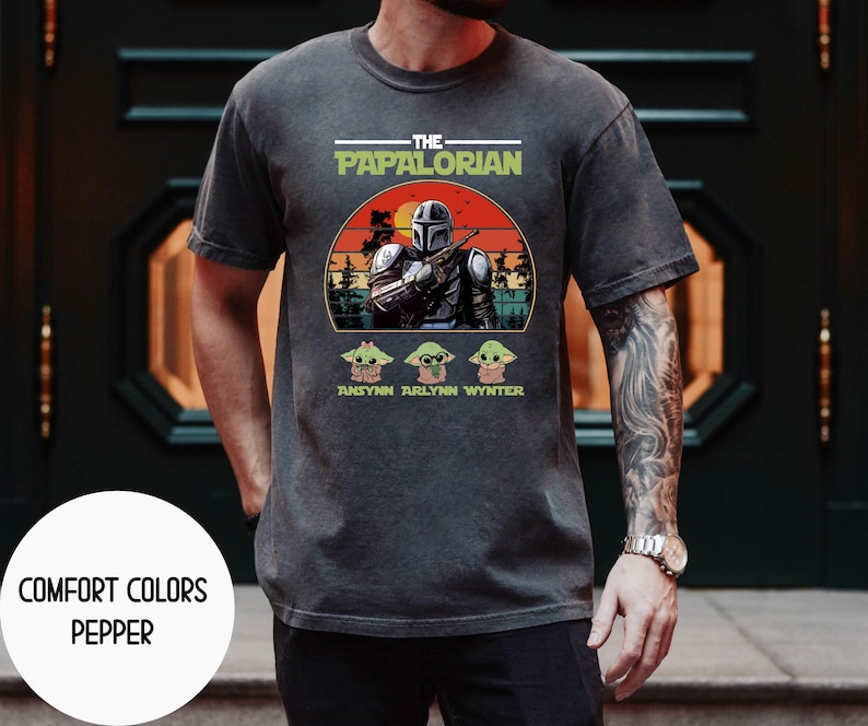 Puede incluir: Camiseta gris oscuro con un gr&aacute;fico que presenta un personaje al estilo Mandaloriano y el texto "THE PAPALORIAN". Debajo del gr&aacute;fico hay tres personajes de Grogu y los nombres "ANSYNN ARLYNN WYNTER". La camiseta tambi&eacute;n tiene el texto "COMFORT COLORS PEPPER".