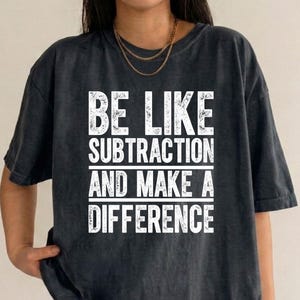 May include: A charcoal gray short-sleeve t-shirt with a distressed white graphic print on the chest. The text reads BE LIKE SUBTRACTION AND MAKE A DIFFERENCE, arranged in five lines with horizontal lines separating the phrases.