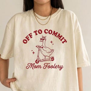 May include: A cream-colored t-shirt featuring a red graphic of a goose wearing heart-shaped sunglasses and a bandana while riding a scooter. The text reads "OFF TO COMMIT" above the goose and "Mom Foolery" below, accented by small star icons.