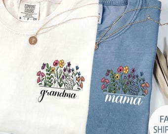 May include: Two t-shirts, one cream and one blue, with floral designs and the words "grandma" and "mama" in script. Each shirt has a gold necklace. The floral design includes pink, yellow, and purple flowers.