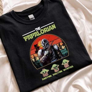 Puede incluir: Camiseta negra con un dise&ntilde;o de The Mandalorian, un atardecer y tres personajes de Grogu. El texto "THE PAPALORIAN" est&aacute; impreso encima del dise&ntilde;o, y "ANSYNN ARLYNN WYNTER" est&aacute; impreso debajo.