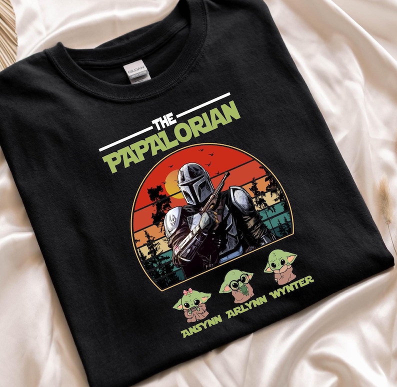 Puede incluir: Camiseta negra con un dise&ntilde;o de The Mandalorian, un atardecer y tres personajes de Grogu. El texto "THE PAPALORIAN" est&aacute; impreso encima del dise&ntilde;o, y "ANSYNN ARLYNN WYNTER" est&aacute; impreso debajo.