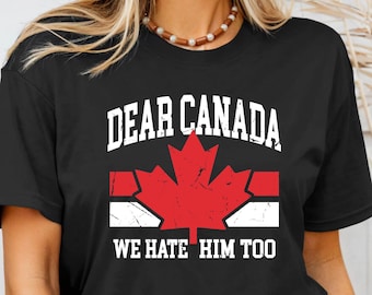 May include: Black t-shirt with the text "DEAR CANADA" above a red maple leaf design and the words "WE HATE HIM TOO" in white. The shirt is a crew neck style.