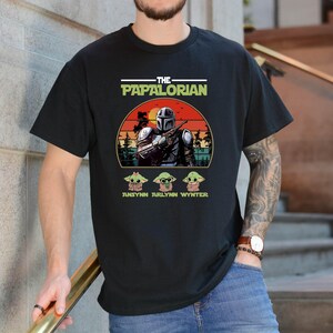 Puede incluir: Camiseta negra con un dise&ntilde;o gr&aacute;fico. El dise&ntilde;o presenta el texto "THE PAPALORIAN" en blanco y verde, una ilustraci&oacute;n de un personaje con casco y tres peque&ntilde;as ilustraciones de personajes. Los nombres "ANSYN ARLYNN WYNTER" est&aacute;n debajo.
