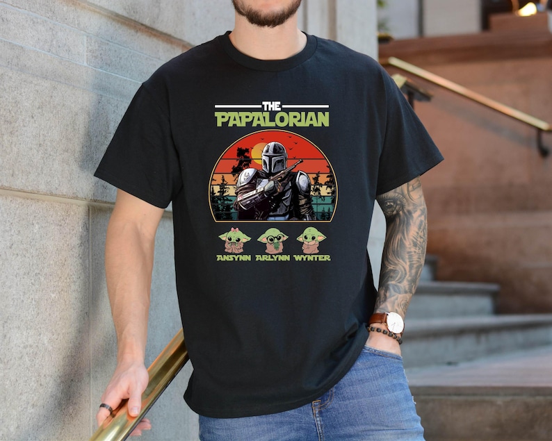 Puede incluir: Camiseta negra con un dise&ntilde;o gr&aacute;fico. El dise&ntilde;o presenta el texto "THE PAPALORIAN" en blanco y verde, una ilustraci&oacute;n de un personaje con casco y tres peque&ntilde;as ilustraciones de personajes. Los nombres "ANSYN ARLYNN WYNTER" est&aacute;n debajo.