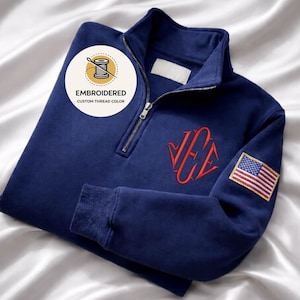 May include: Navy blue quarter-zip sweatshirt with red embroidered monogram and an American flag patch on the sleeve. The sweatshirt features a zipper closure and ribbed cuffs. The text "EMBROIDERED CUSTOM THREAD COLOR" is displayed on a circular logo.