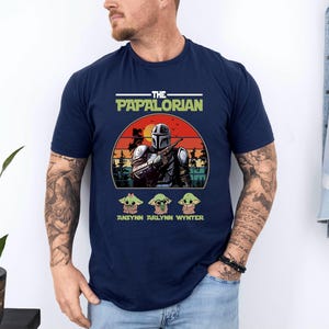 Puede incluir: Camiseta azul marino con un gr&aacute;fico de un personaje al estilo Mandaloriano, el texto "THE PAPALORIAN" y tres personajes Grogu. El dise&ntilde;o est&aacute; ambientado sobre un fondo de puesta de sol. Debajo de los personajes Grogu hay tres nombres.