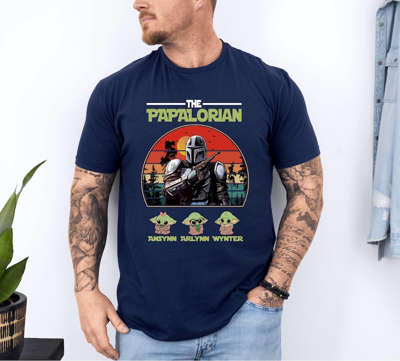 Puede incluir: Camiseta azul marino con un gr&aacute;fico de un personaje al estilo Mandaloriano, el texto "THE PAPALORIAN" y tres personajes Grogu. El dise&ntilde;o est&aacute; ambientado sobre un fondo de puesta de sol. Debajo de los personajes Grogu hay tres nombres.