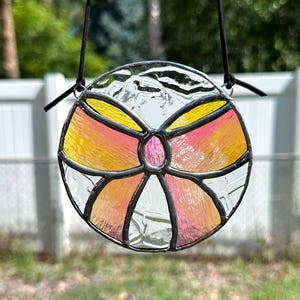 May include: A round stained glass suncatcher with a black metal frame. The design features a central pink oval surrounded by yellow and pink sections, with clear glass accents. A black cord is attached for hanging.
