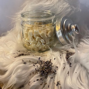 May include: A clear glass jar filled with small, off-white cubes, with the lid slightly ajar. The jar sits on a fluffy, white surface, with dried lavender scattered around it. The jar has a silver lid.