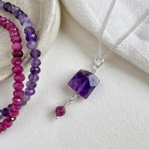 Peut inclure: Un collier en argent avec un pendentif en améthyste violette facettée et une petite pierre précieuse rouge ronde. Un bracelet de perles avec des pierres précieuses violettes et rouges est visible en arrière-plan. Les bijoux sont présentés sur une surface blanche.