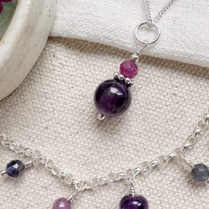 Puede incluir: Un collar de plata con un gran colgante de amatista púrpura oscuro y una piedra preciosa rosa más pequeña encima. La cadena tiene dijes adicionales de piedras preciosas púrpuras y azules.