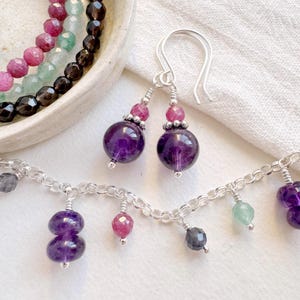 May include: A selection of gemstone jewellery, including a necklace and earrings. The earrings feature large, round purple amethyst beads with smaller pink and silver accents. The necklace has a silver chain with various gemstone charms.