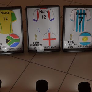 May include: Three framed prints featuring football jerseys with the text Name and 12 or 7. Each print displays a FIFA World Cup 2026 logo, a country flag, and the country name: South Africa, England, and Argentina. The frames are black and rectangular.