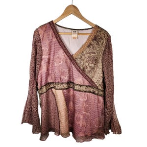 May include: A long-sleeved V-neck blouse featuring a patchwork design with various floral and geometric patterns in shades of mauve, brown, and cream. The garment has a wrap-style front, a defined waist panel, and flared sleeves, all displayed on a wooden hanger.