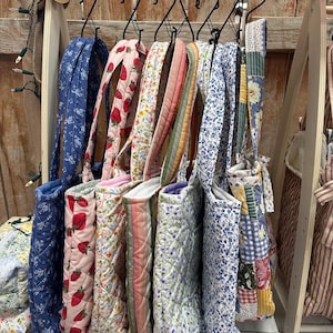 May include: A collection of quilted tote bags hanging on a wooden rack. The bags feature various patterns including blue floral, red strawberry print, small multicolored flowers, striped fabric, purple floral, and a patchwork design with squares.
