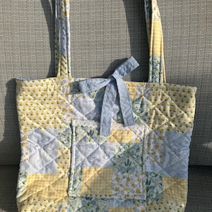 May include: A quilted tote bag with a patchwork design in yellow, blue, and white floral and gingham patterns. The bag features a light blue bow and a pocket with lace trim. The handles are made of the same patchwork fabric.