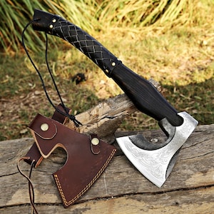 May include: A hand-forged Viking axe with a black leather wrapped handle and a brown leather sheath. The axe head has a detailed etched design.