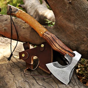 May include: A handcrafted axe with a wooden handle and a leather-wrapped grip. The axe head is silver with etched designs. A brown leather sheath protects the blade. The axe is displayed on a weathered wooden surface.