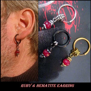 Può includere: Un paio di orecchini con un cerchio d'argento e oro e una pietra preziosa rossa pendente. Gli orecchini sono etichettati "Ruby & Hematite Earring".
