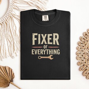 Könnte beinhalten: Schwarzes T-Shirt mit dem Text "Fixer of Everything" in braunen und weißen Buchstaben. Unter dem Text ist ein Schraubenschlüssel abgebildet.