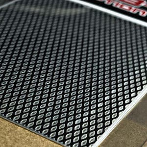 Puede incluir: Primer plano de una superficie negra y plateada con un patrón. El patrón consiste en una serie repetida de diseños en forma de diamante. La superficie tiene un acabado brillante y está bordeada por un borde plateado. La imagen está bien iluminada, resaltando la textura.
