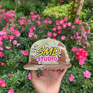 Puede incluir: Una gorra de béisbol con estampado de camuflaje en tonos beige, marrón y negro con visera plana. El frente presenta un parche bordado con el texto amarillo SMD delineado en blanco sobre el texto rosa STUDIO, también delineado en blanco.