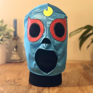May include: A blue wrestling mask with red and yellow accents. The mask has a large mouth opening and two eye holes.