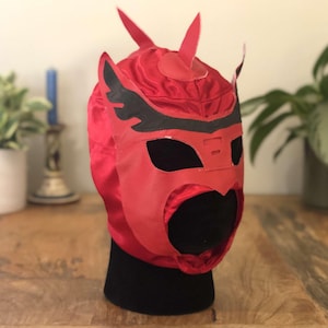 Puede incluir: Máscara de lucha libre roja con detalles negros y una parte superior puntiaguda. La máscara tiene una gran abertura para los ojos y una abertura más pequeña para la boca.