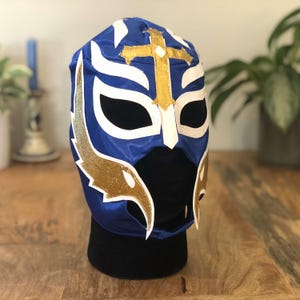 May include: Blue wrestling mask with a white and gold design. The mask has a cross on the forehead and a black eye mask.