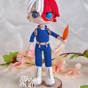 May include: Crocheted doll of a character with white and red hair, blue jumpsuit, and white boots. The doll has two different colored eyes, one gray and one blue. The doll is holding an orange object. The doll is standing on a wooden base with the text "Tinna Rumi".