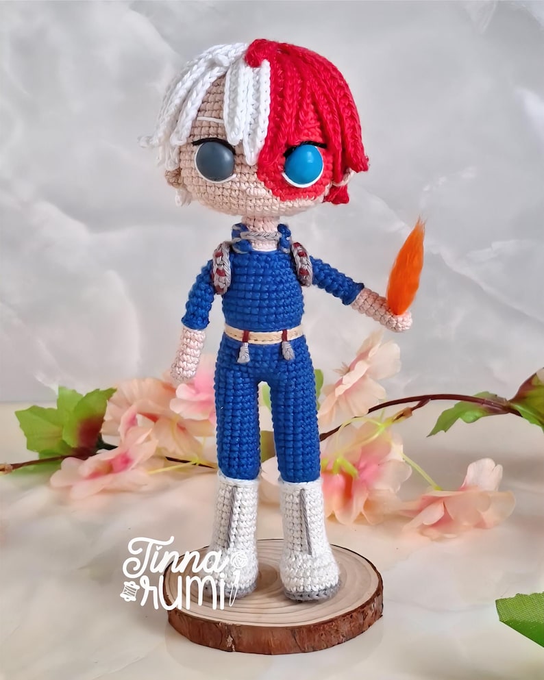 May include: Crocheted doll of a character with white and red hair, blue jumpsuit, and white boots. The doll has two different colored eyes, one gray and one blue. The doll is holding an orange object. The doll is standing on a wooden base with the text "Tinna Rumi".