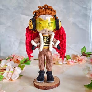May include: A crocheted doll depicting a character with red wings, headphones, and a yellow visor. The doll is dressed in a brown jacket with white trim, brown trousers, and black boots. It is displayed on a wooden base, highlighting the detailed crochet work and bright colours.