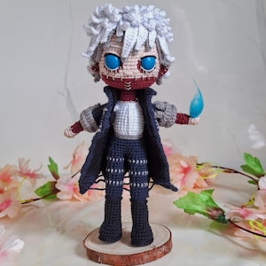 May include: Crocheted doll of a character with white hair, blue eyes, and a red stitched face. The doll wears a dark blue coat, pants, and gray gloves. It stands on a wooden base and holds a blue flame. The doll is surrounded by pink flowers.