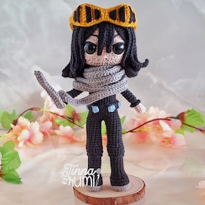 May include: A handcrafted crochet doll depicting a character with black hair, a grey scarf, and yellow goggles. The doll wears a black jumpsuit with blue details and stands on a wooden base. It has large, black eyes and holds a grey object.