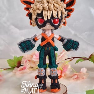 May include: A handmade crochet doll of a character with spiky blonde hair, red eyes, and a black mask. The doll wears a black and orange outfit with green accents, gray gloves, and knee pads. It stands on a wooden slice, with the artist's signature visible.